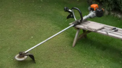 Débroussailleuse thermique posée dans l'herbe tondue, appuyée sur un banc de jardin avec un chiffon et des outils